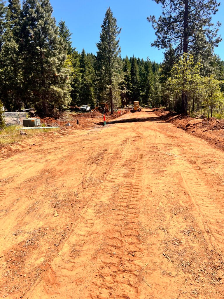 Excavation crew grading a dirt road through a forested Southern Oregon property with heavy equipment and fresh track marks visible on the surface