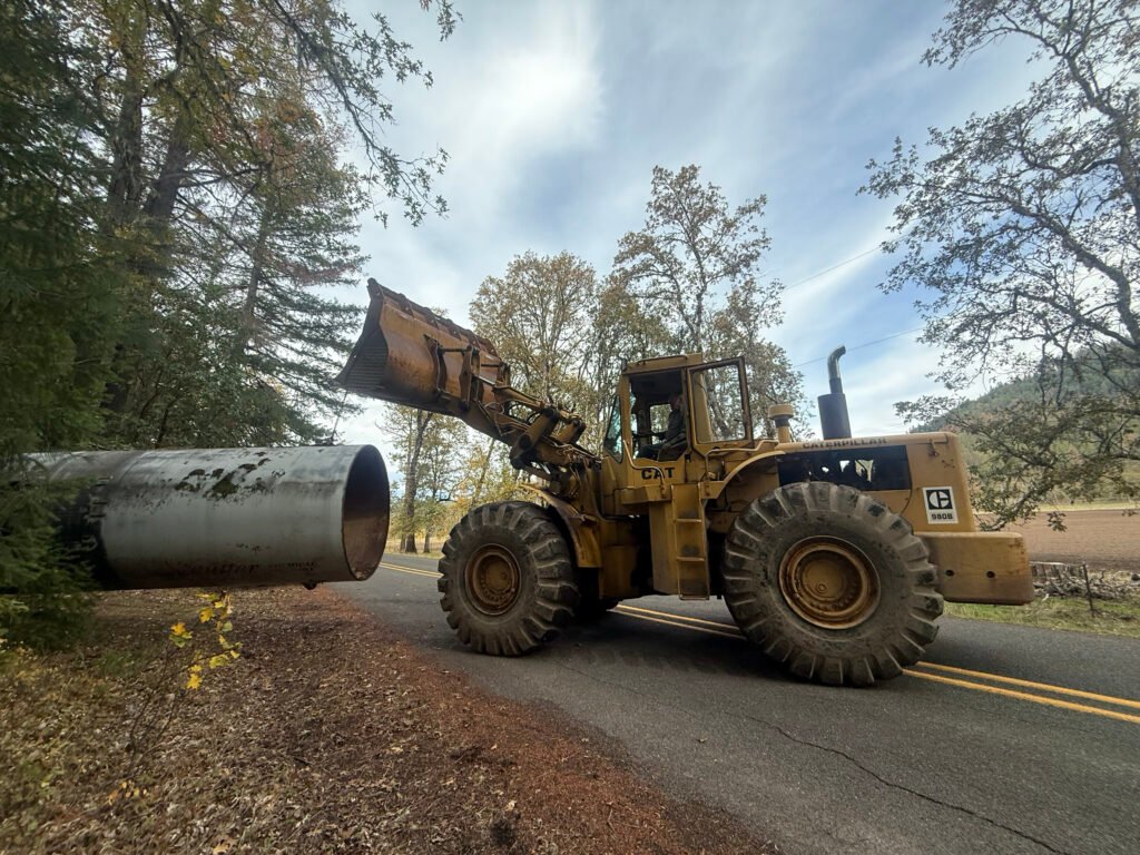 Front loader placing a large metal culvert pipe along a rural roadside for drainage installation and site access work